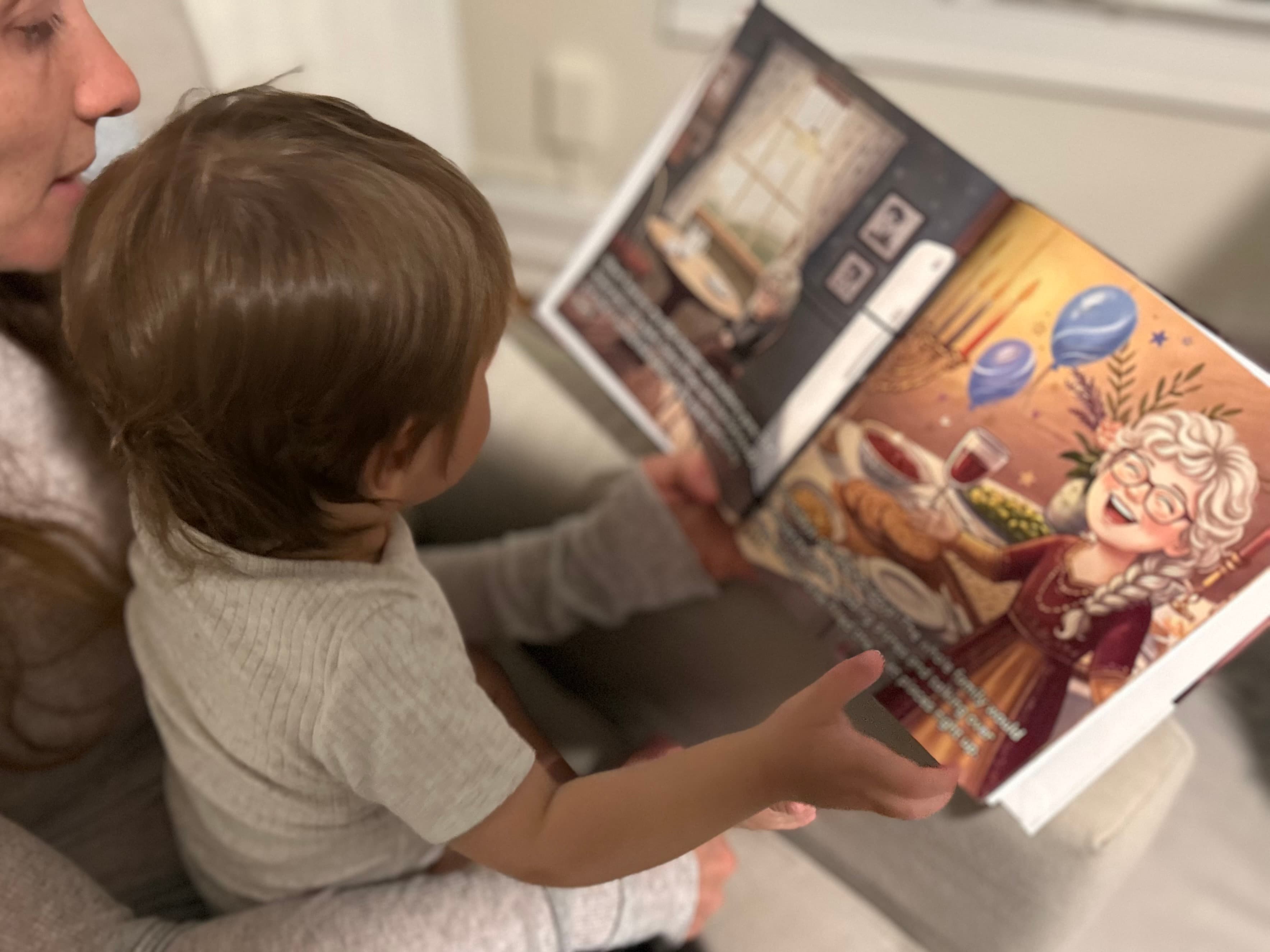 A mother holding her toddler in her lap as they read a Family Hero Book together. An open spread shows an illustrated grandmother in her kitchen.